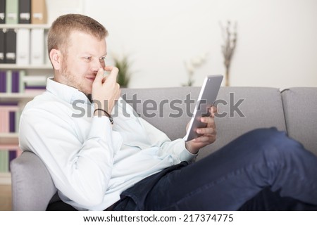Man snickering to himself as he reads his tablet computer holding his hand in front of his face as he lies back comfortably on a sofa at home