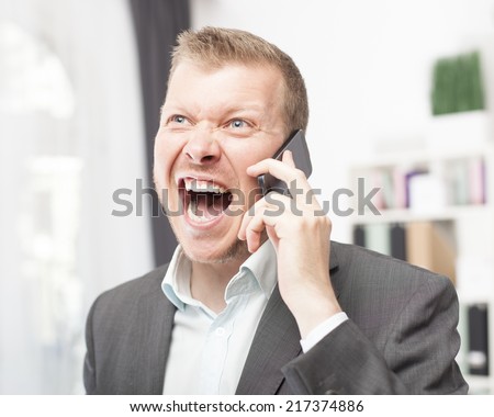 Exuberant young man in a suit jacket and open neck shirt shouting in reaction to a call on his mobile phone as he stands indoors at the office