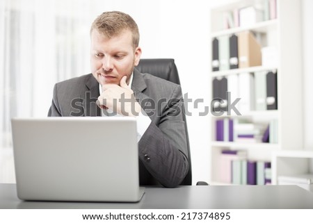Businessman sitting at his desk in the office looking at his laptop with a wry expression grimacing as he reads information on the screen