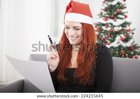 young red hair girl writing a wish list and wearing a red santa hat in front of a firtree with lights and balls