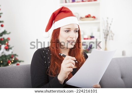young red hair girl writing a wish list and wearing a red santa hat in front of a firtree with lights and balls, sitting on a sofa/ couch