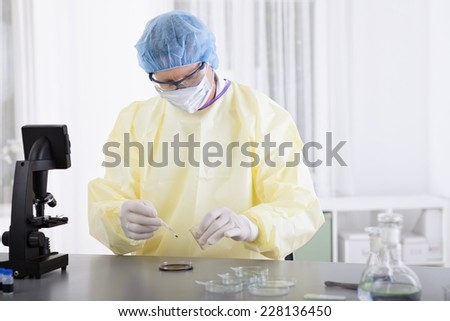 Scientist in protective gear holding blood sample with petri dish and microscope foreground in laboratory