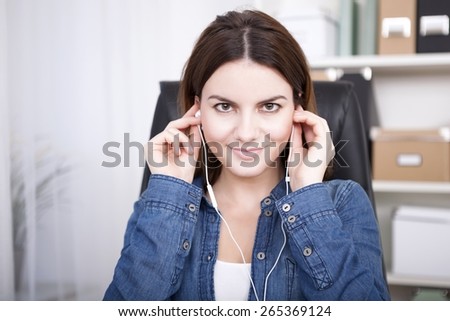 Businesswoman sitting listening to music or a recording on a set of earplugs looking directly into the lens with a warm friendly smile