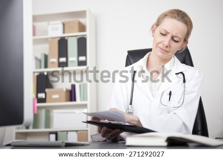 Adult Woman Physician Scanning Medical Reports on a Clipping Board While Sitting at her Worktable.