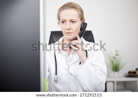 Close up Female Physician Calling Through Telephone and Looking at the Computer Monitor Seriously While Sitting at her Worktable.