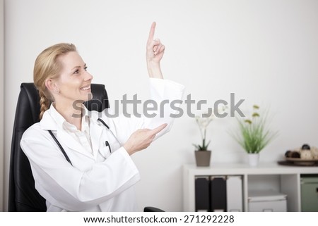 Smiling Woman Physician Pointing and Looking to Upper Right Side with Copy Space While Sitting at her Office.