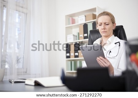 Serious Adult Female Medical Doctor Reading at her Tablet Computer While Sitting at her Desk.