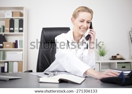 Happy Adult Woman Physician in White Medical Gown Leaning on her Table While Talking to Someone Over the Phone