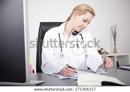Serious Blond Woman Medical Doctor Talking on Telephone While Writing at her Office.