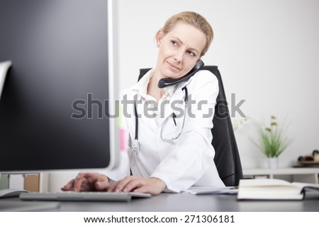 Serious Adult Female Doctor Talking on Telephone and Using her Desktop Computer While Sitting at her Office.