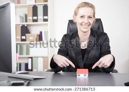 Close up Smiling Businesswoman Pointing at Miniature Model House on Top of her Desk While Looking at the Camera.
