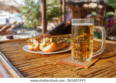 Natural light photo with shallow DOF of chicken baguette on plate with glass of beer on mat on bamboo table with cafe background