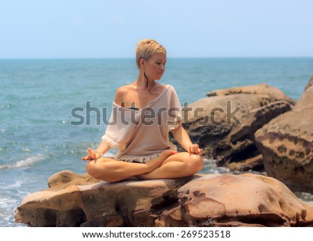 Beautiful woman seated in yoga pose at rocky blue sea background horizontal view