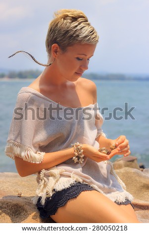 Beautiful young woman sitting on picturesque rocky seashore playing with shells