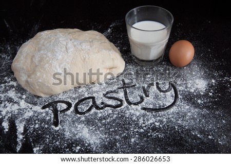 Closeup of pastry dough and egg with milk on black marble table with handwritten word on scattered flour
