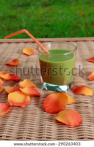 Blended green smoothie and colorful flower petals on rattan table with green grass at background