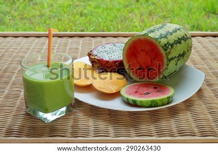Blended green smoothie and sliced watermelon, mango and dragon fruit on white plate on rattan table, horizontal view 2