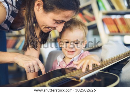Young boy is trying to play guitar, mother is watching him