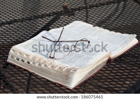 A closeup of a pair of reading glasses resting on top of a worn, open Bible on a wrought-iron patio table