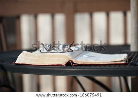 A pair of reading glasses rests on top of an open Bible on a wrought-iron patio table on a wooden deck as the evening shadows settle in.