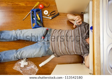 A man on the kitchen floor is fixing the sink.
