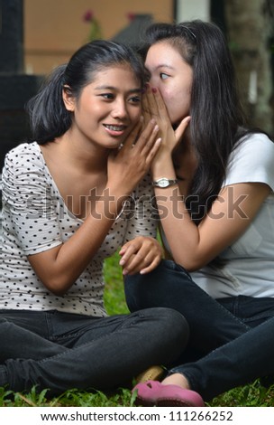 Two pretty southeast asian girls sharing exciting secret stories / gossiping  with happy expression at outdoor scene
