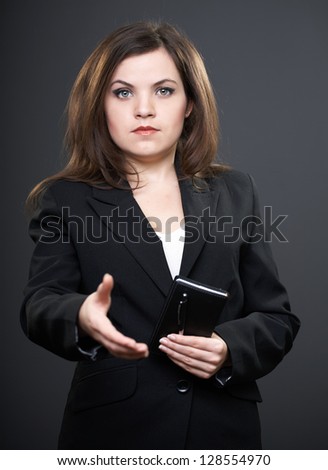 Attractive young woman in a black jacket. Woman holds a notebook and a pen. Woman gives a hand to shake. On a gray background