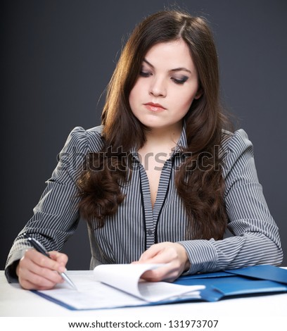 Attractive young woman in a gray blouse sitting at the table. Woman signing documents. On a gray background