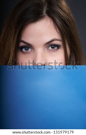 Attractive young woman in a gray blouse. Woman holds a blue folder and covers her face. On a gray background