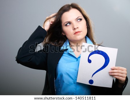 Attractive young woman in a black jacket. Woman holds a poster with a big question mark. Thinking and looking to the left. On a gray background