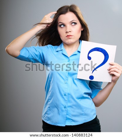 Attractive young woman in a blue shirt. Woman thinks and holds poster with big question mark. Isolated on gray background