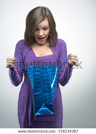 Surprised young woman in a lilac dress. Holding blue gift bag. On a gray background