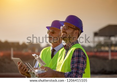 Two engineers with helmets and vests operating with drone by remote control and looking up in the sky. Technology innovations in construction industry