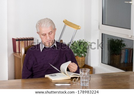 Old man with crutches behind reading book at table