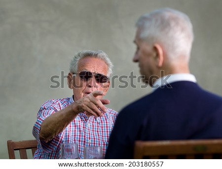 Old man with black sunglasses pointing forefinger in another old man