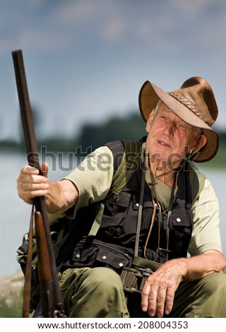 Senior hunter with shotgun sitting beside lake