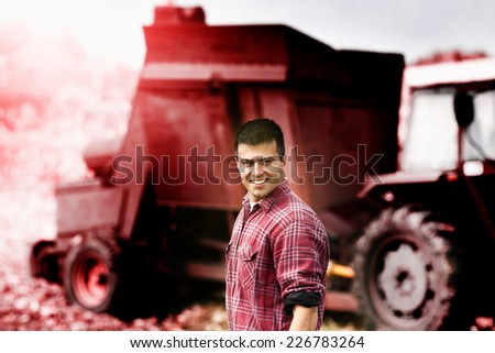 Young satisfied farmer standing in front of agricultural machinery in corn field