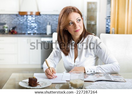 Middle aged woman sitting at dining table with recipe book, cake in plate in front of her and thinking about something nice