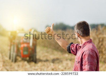 Young satisfied farmer showing thumbs up, tractor on field in background