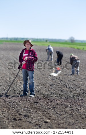 Senior peasant with hoe standing on fertile land, other peasants hoeing and sowing in background