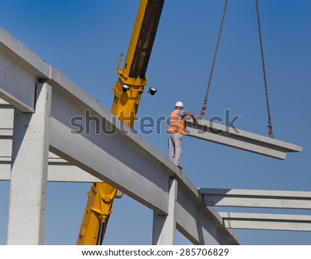 Construction worker standing on concrete beam on height and placing truss lifted by crane