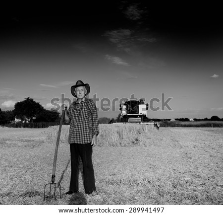 Old farmer with hat and hayfork standing on field during harvest, combine harvester in background, black and white image
