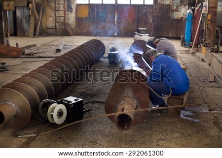 Two workers welding ship part in messy workshop