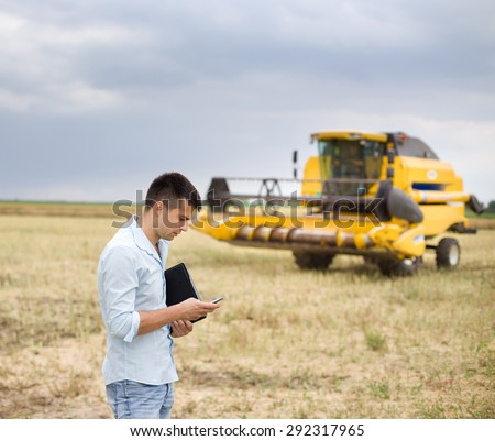 Young businessman standing with laptop and cell phone on field, combine harvester in background
