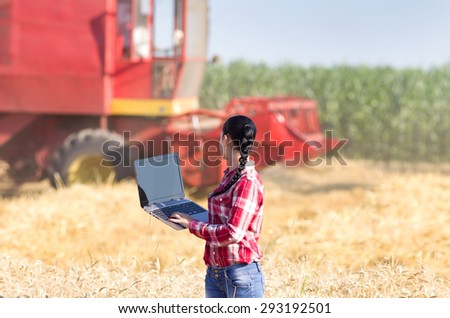 Young beautiful woman in plaid shirt standing with laptop in wheat field with combine harvester in background