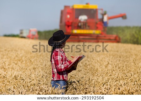 Young beautiful woman in plaid shirt and with hat writing notes in wheat field during harvest, combine harvester in background