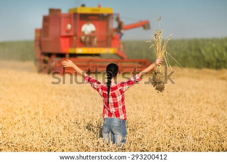 Young woman in plaid shirt standing on wheat field with raised arms in front of combine harvester