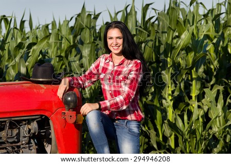 Portrait of smiling young woman with black long hair in plaid shirt sitting on tractor in corn field