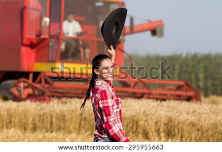 Pretty cowgirl with hat in raised arm looking over shoulder in camera in the golden wheat field, combine harvester in background