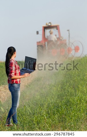 Young woman with laptop standing in the field and looking at tractor with hay rake